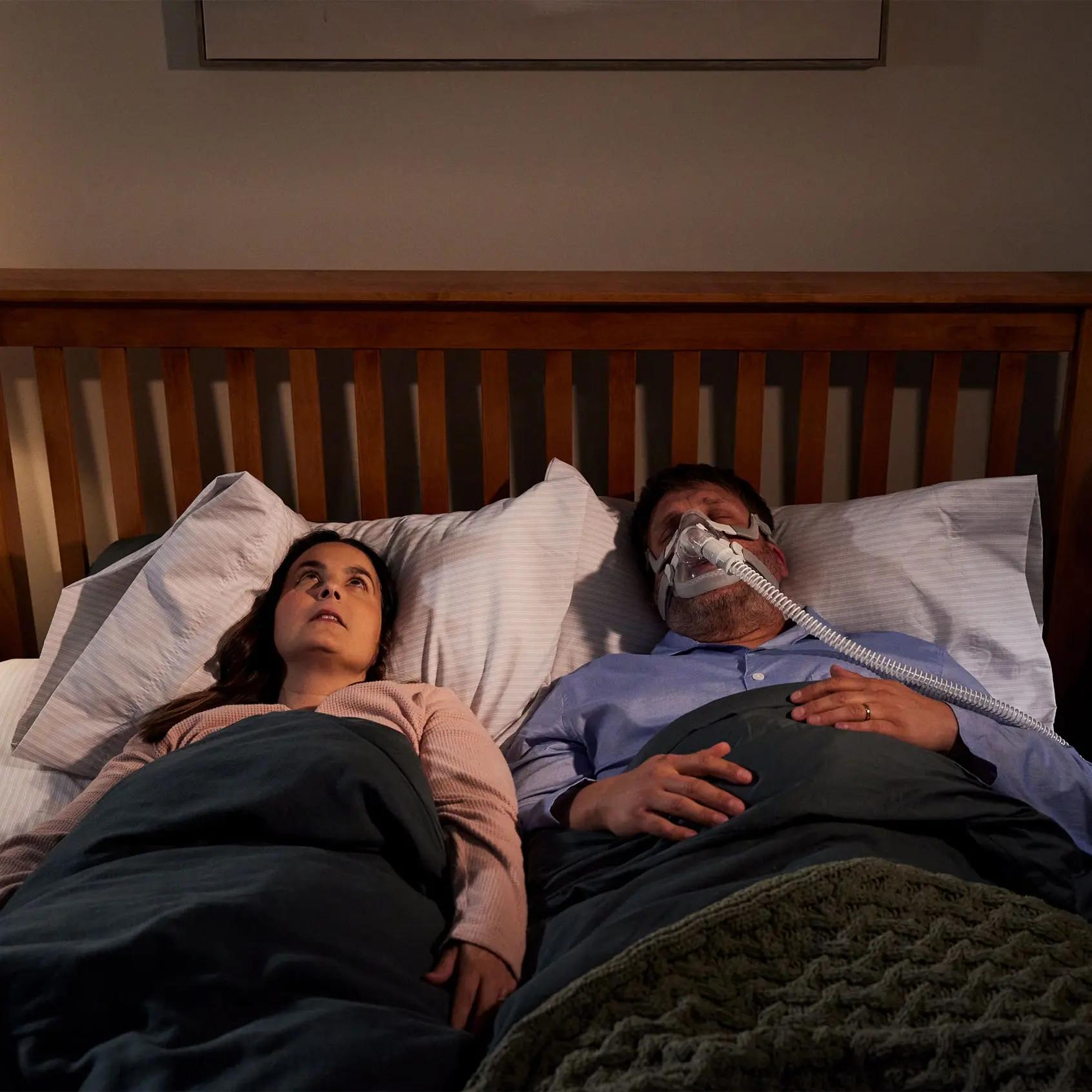 The image depicts a man and a woman lying side by side in bed. The man is wearing a CPAP mask, the woman looks frustrated.